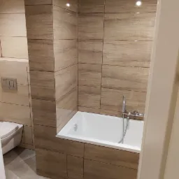 Nowoczesna łazienka z wanną obudowaną płytkami imitującymi drewno, marmurowa podłoga, wisząca toaleta i minimalistyczna armatura.