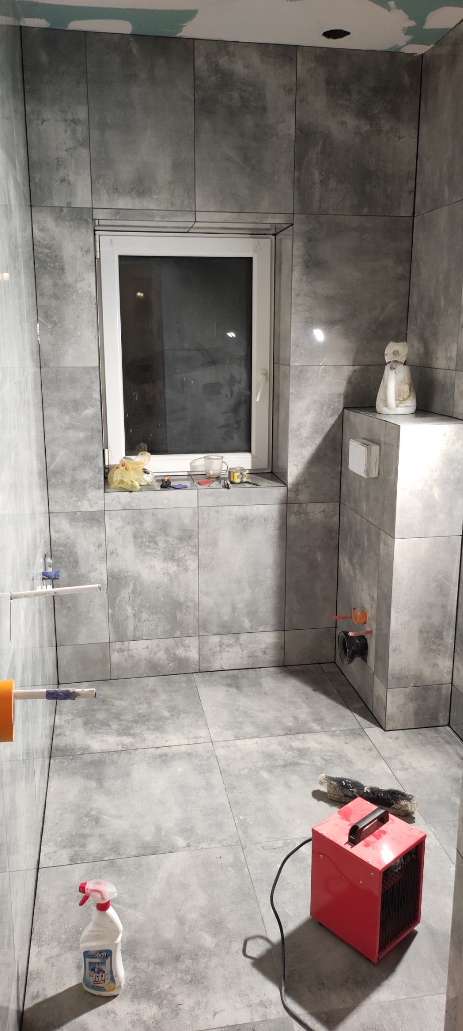 Szara łazienka w trakcie prac wykończeniowych, widoczne płytki na ścianach i podłodze, okno z narzędziami na parapecie, grzejnik elektryczny i butelka środka czyszczącego na podłodze.