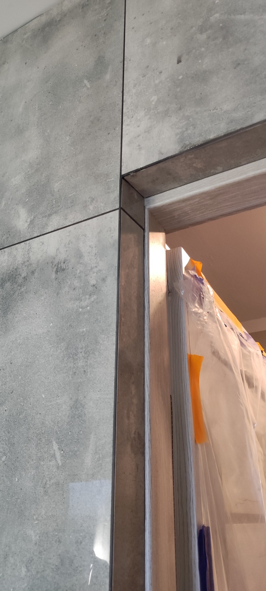 Fragment łazienki w trakcie prac: szare płytki imitujące beton na ścianie, widoczna ościeżnica drzwi zabezpieczona folią malarską z pomarańczową taśmą.