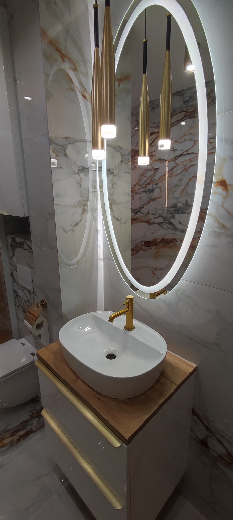 Nowoczesna łazienka z owalną umywalką nablatową, złotą armaturą i podświetlanym lustrem z marmurowym tłem oraz wiszącymi lampami.