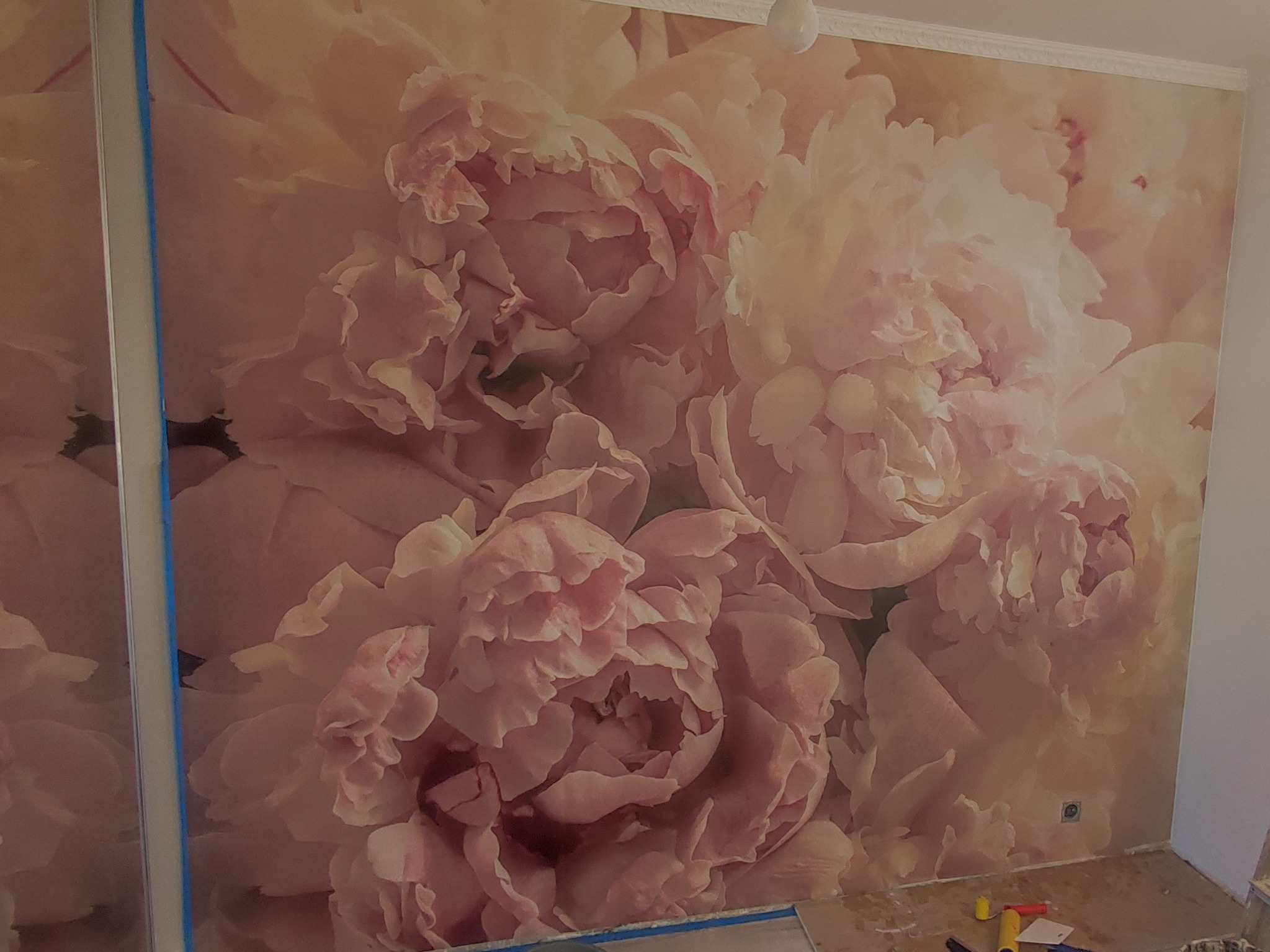 Ściana w pokoju pokryta fototapetą z motywem dużych, różowych piwonii, widoczne narzędzia malarskie i resztki kleju na podłodze.