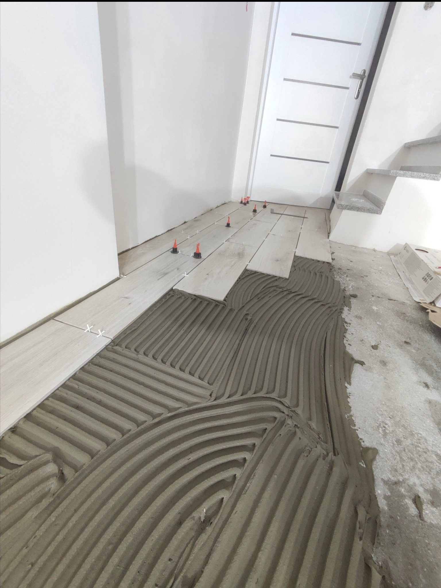 Proces układania podłogi z płytek imitujących drewno, widoczna warstwa kleju z wyżłobieniami, system poziomowania z czerwonymi klinami, białe krzyżyki dystansowe, białe ściany i drzwi w tle.