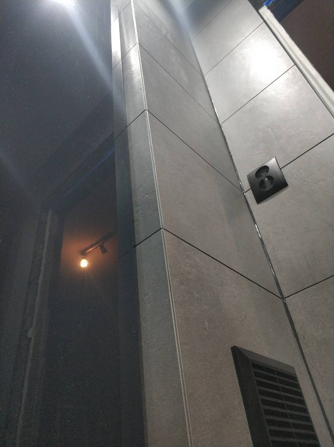 Ściana łazienki wyłożona szarymi płytkami ceramicznymi, widoczny czarny włącznik światła i kratka wentylacyjna, z odbiciem lampy w lustrzanej powierzchni.