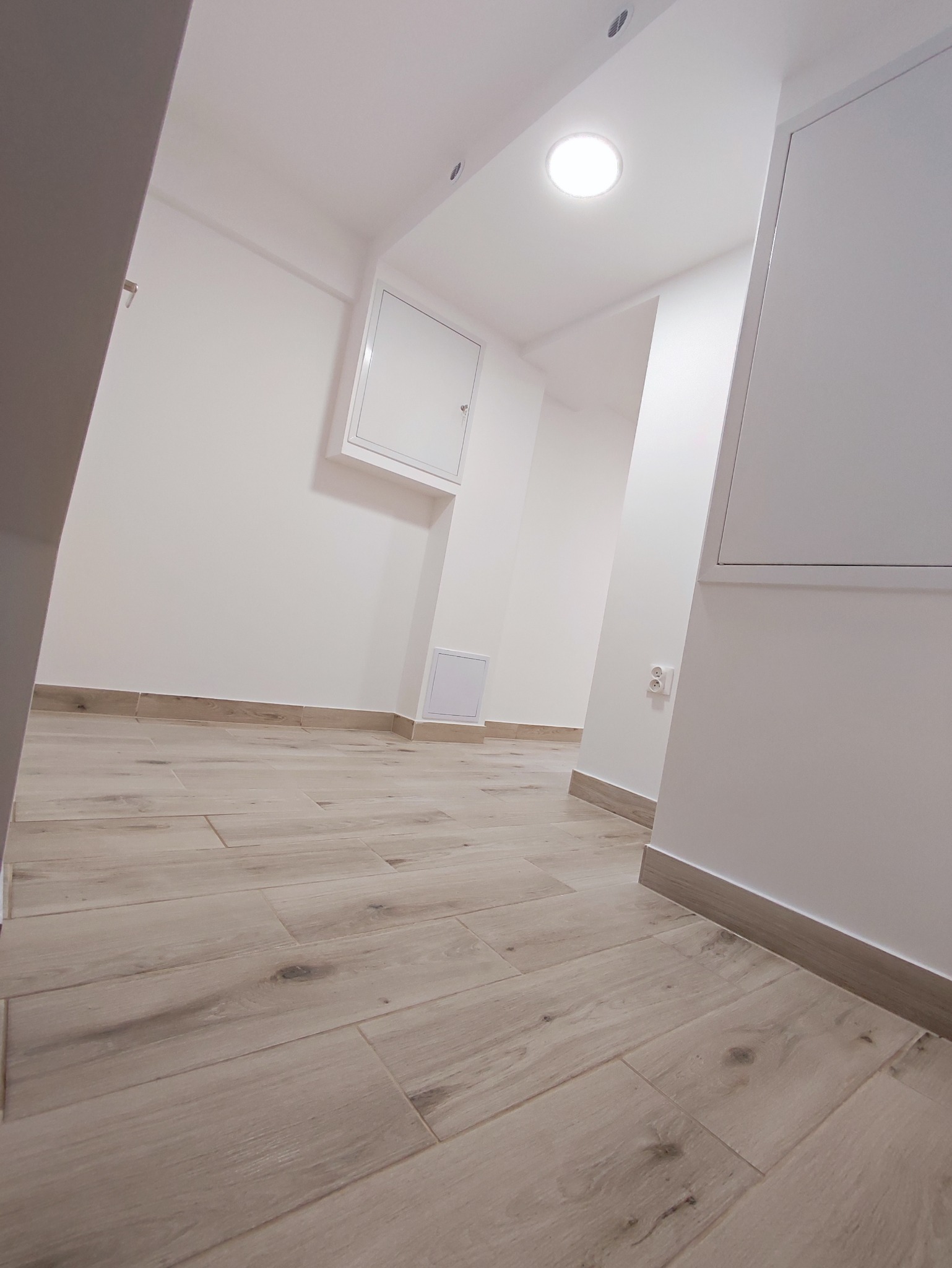 Jasne płytki podłogowe imitujące drewno w korytarzu z białymi ścianami, widoczne włazy rewizyjne i gniazdko elektryczne.