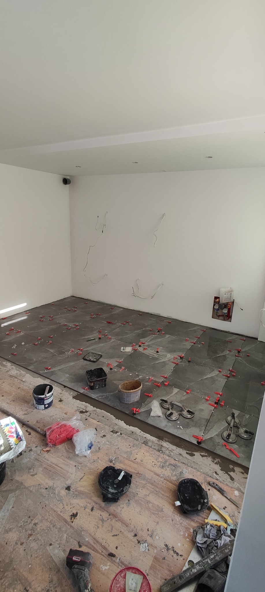 Układanie szarych płytek podłogowych w pomieszczeniu z białymi ścianami, widoczne narzędzia glazurnicze, krzyżyki dystansowe i przyssawki do płytek.