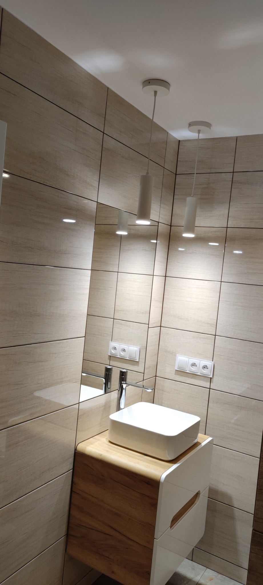 Nowoczesna łazienka z beżowymi, drewnopodobnymi płytkami, białą umywalką nablatową na szafce z drewnianym blatem i białymi frontami, oświetlona cylindrycznymi lampami wiszącymi.