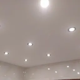Biały sufit podwieszany z równomiernie rozmieszczonymi, okrągłymi lampami LED, odbijającymi się w błyszczącej powierzchni płytek ściennych.