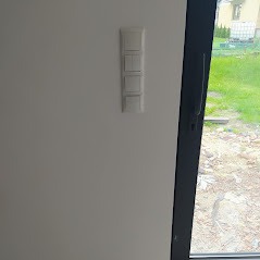 Biały, poczwórny włącznik światła na jasnej ścianie w nowym budynku, widoczny fragment okna z widokiem na zewnątrz.