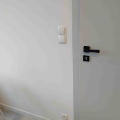 Biała ściana z podwójnym, białym włącznikiem światła i szarą, matową ścianą obok białych drzwi z czarną klamką i zamkiem.