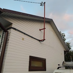 Zewnętrzna instalacja odgromowa na ścianie domu z sidingu, widoczna rura spustowa w kolorze czerwonym i przewody elektryczne prowadzone po elewacji.