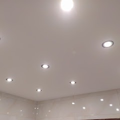 Biały sufit podwieszany z równomiernie rozmieszczonymi, okrągłymi lampami halogenowymi, odbijającymi światło na błyszczących płytkach ściennych.