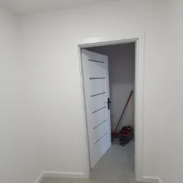 Otwarta biała, minimalistyczna drzwi wewnętrzne z poziomymi frezowaniami, widok na szare płytki podłogowe i akcesoria do sprzątania w rogu pomieszczenia, ściany pomalowane na biało.
