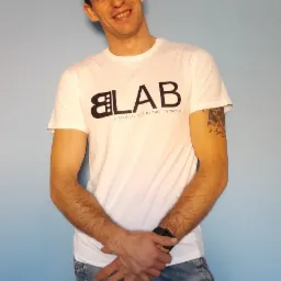 Uśmiechnięty mężczyzna w białej koszulce z logo 'LAB' i napisem 'przeszkody OCR Bartosz Szymański', stojący na tle błękitnej ściany.