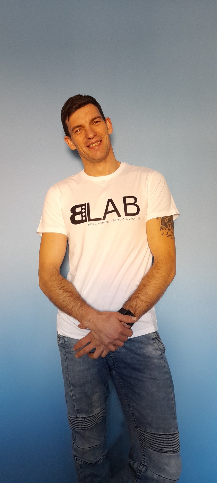Uśmiechnięty mężczyzna w białej koszulce z logo 'LAB' i napisem 'przeszkody OCR Bartosz Szymański', stojący na tle błękitnej ściany.