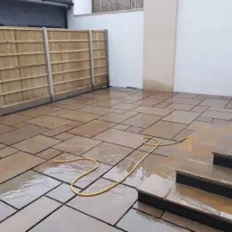 Świeżo ułożony, mokry taras z nieregularnych płyt kamiennych w odcieniach brązu, z niskimi schodami i jasnym, drewnianym ogrodzeniem w tle, zwinięty wąż ogrodowy leży na powierzchni.