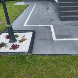 Nowoczesny taras wyłożony szarą płytą z dekoracyjną obwódką z białych kamyków, z lampą ogrodową otoczoną rabatą z białym grysem i roślinami.