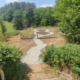 Ścieżka z jasnego kruszywa prowadząca do metalowej bramki i rabatki w kształcie ósemki, otoczona siatką i młodymi roślinami, widok z góry ze schodów wykończonych kostką.