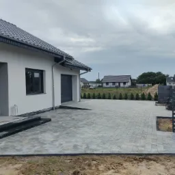 Nowoczesny podjazd z szarej kostki brukowej ułożonej w regularny wzór, prowadzący do garażu domu jednorodzinnego z szarymi oknami i dachem, w tle widoczne inne domy i pochmurne niebo.