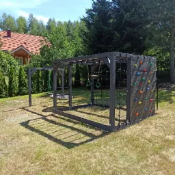 Drewniany plac zabaw w odcieniach szarości z drabinkami, ścianką wspinaczkową z kolorowymi uchwytami i huśtawką, umieszczony na trawniku przed domem z czerwoną dachówką.