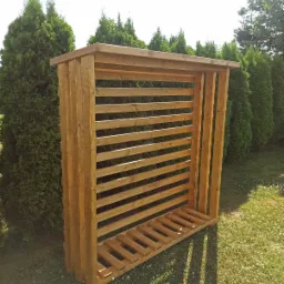 Drewniana konstrukcja z poziomych listew, prawdopodobnie drewutnia, ustawiona na trawniku z kwitnącą koniczyną, z tujami w tle.