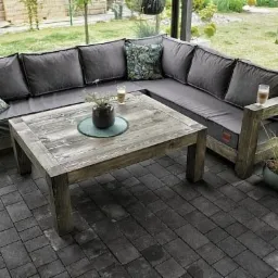 Drewniany narożnik z szarymi poduchami i stolik kawowy na tarasie z kostki brukowej, dwie szklanki i doniczka z rośliną na stole, rośliny w donicach obok narożnika.