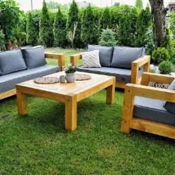 Zestaw mebli ogrodowych z jasnego drewna z szarymi poduszkami na trawniku, w tle żywopłot z tujami.