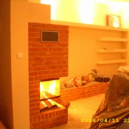 Murowany kominek z cegły z paleniskiem, w trakcie prac wykończeniowych, widoczne narzędzia i materiały budowlane w otoczeniu, oświetlenie ciepłym światłem.