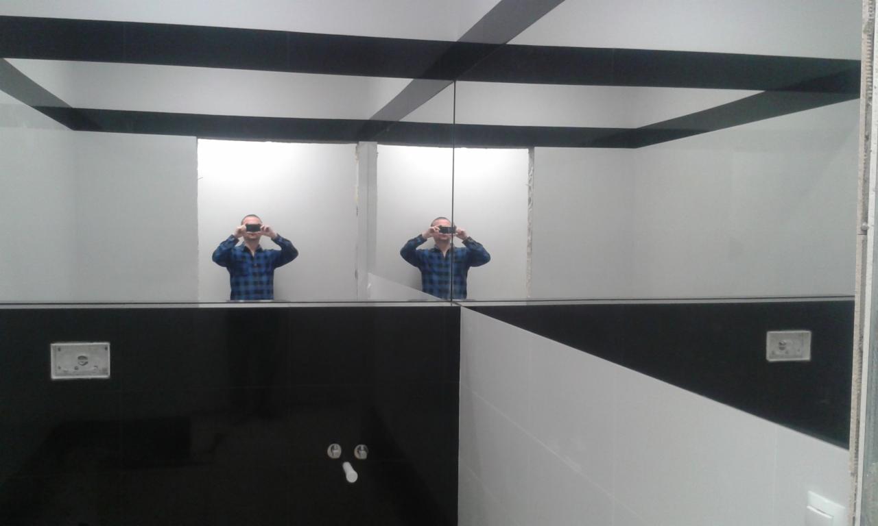 Nowoczesna łazienka z czarno-białymi płytkami ułożonymi w geometryczny wzór, duża tafla lustra odbija osobę robiącą zdjęcie, widoczne elementy instalacji.