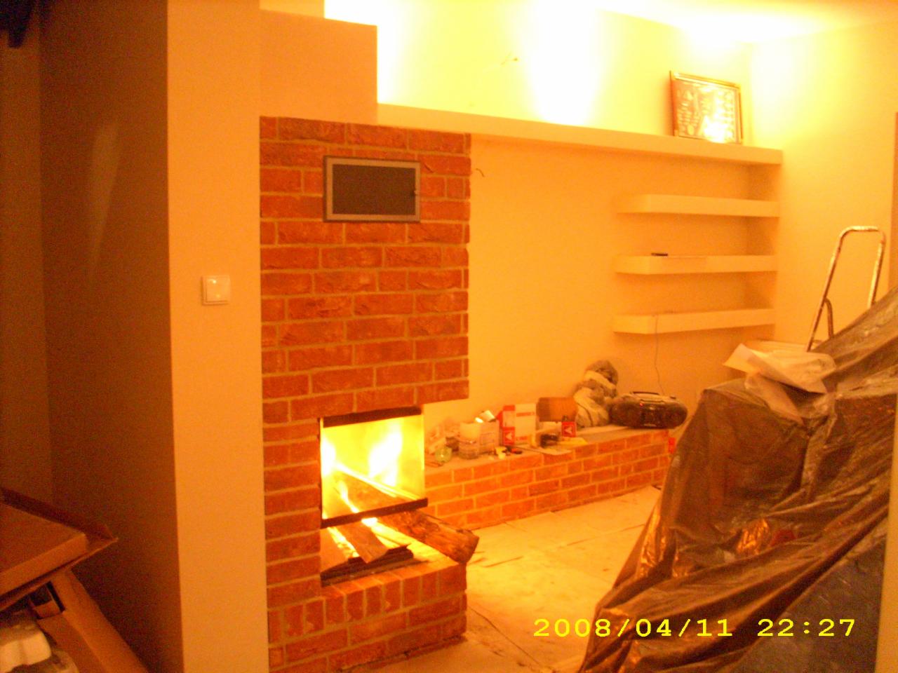 Murowany kominek z cegły z paleniskiem, w trakcie prac wykończeniowych, widoczne narzędzia i materiały budowlane w otoczeniu, oświetlenie ciepłym światłem.