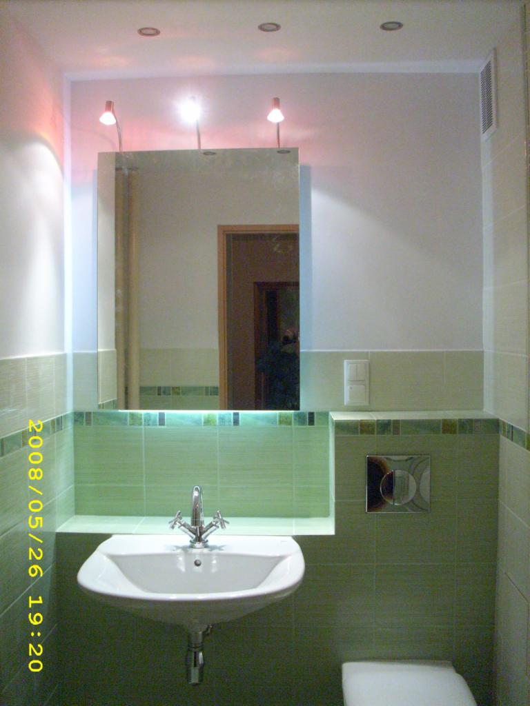 Umywalka wisząca z chromowaną baterią w łazience wykończonej zielonymi płytkami z dekoracyjnym paskiem, nad umywalką lustro z oświetleniem punktowym, widoczny fragment toalety.