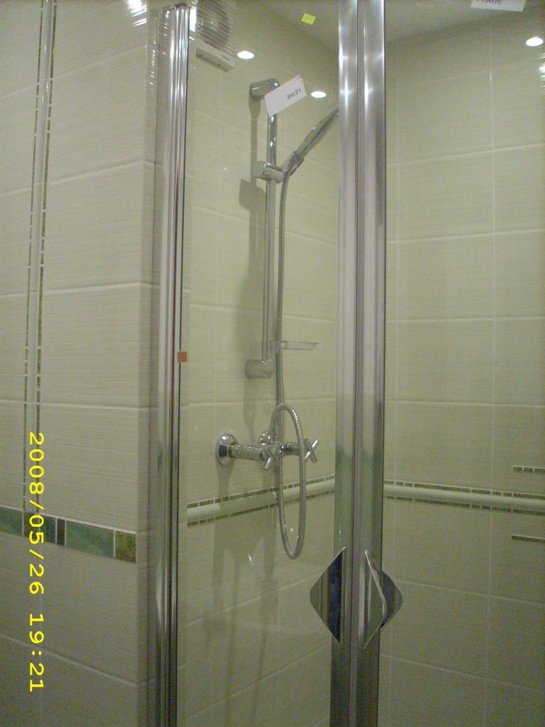 Kabina prysznicowa z chromowanymi elementami, wykończona jasnymi płytkami z zielonym akcentem, widoczne bateria i słuchawka prysznicowa, uchwyt kabiny w kształcie rombu.