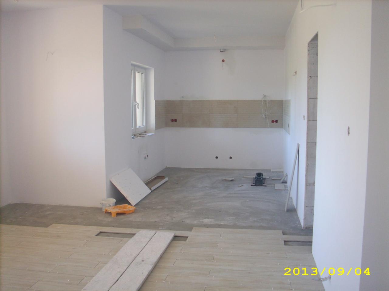 Wnętrze kuchni w trakcie remontu: położone częściowo płytki podłogowe imitujące drewno, fragment ułożonej glazury na ścianie, widoczne narzędzia i materiały budowlane, ściany przygotowane...