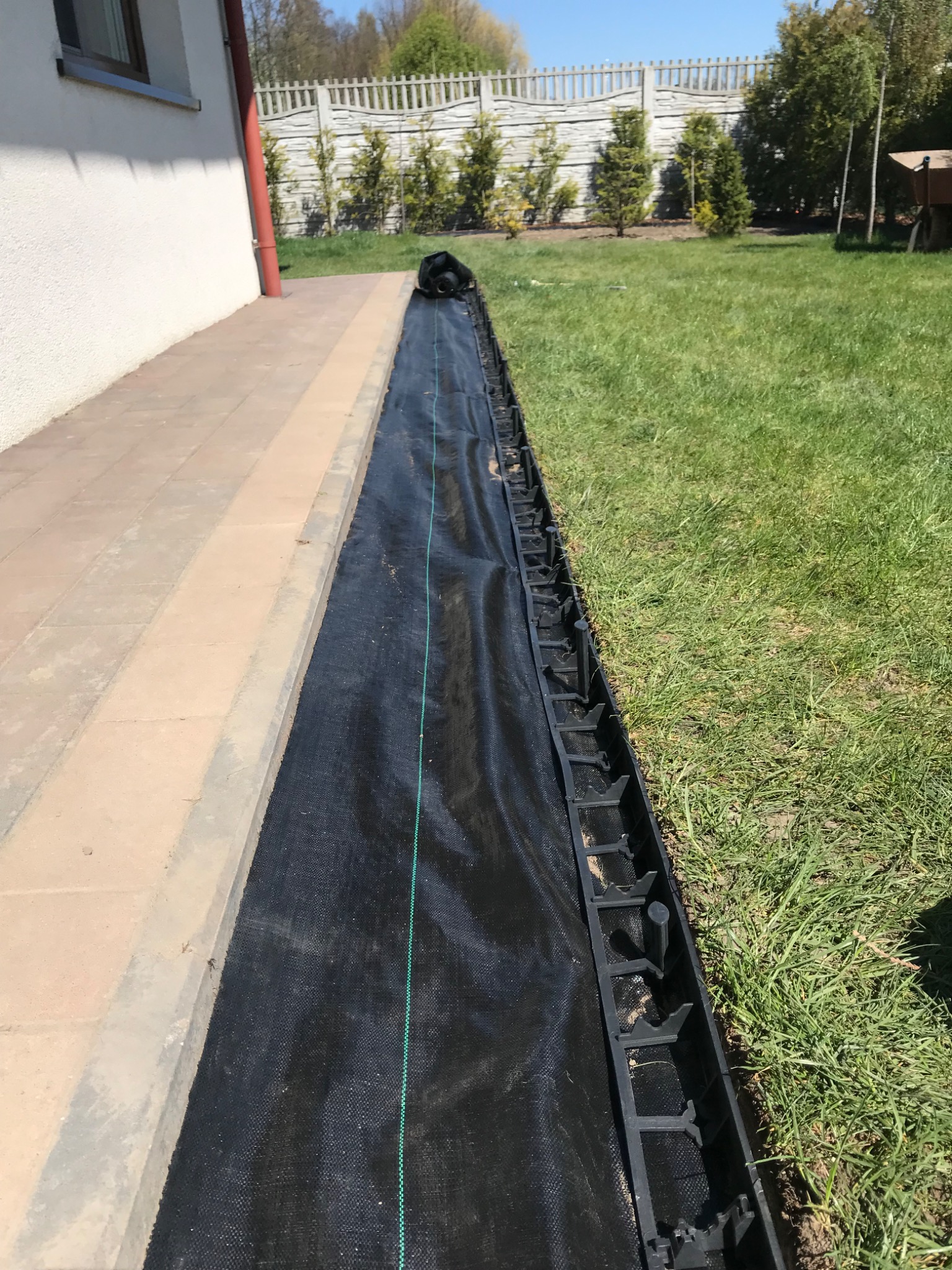 Czarna agrowłóknina rozłożona wzdłuż chodnika, zabezpieczona plastikową obrzeżem trawnikowym, widoczna trawa i roślinność w tle.