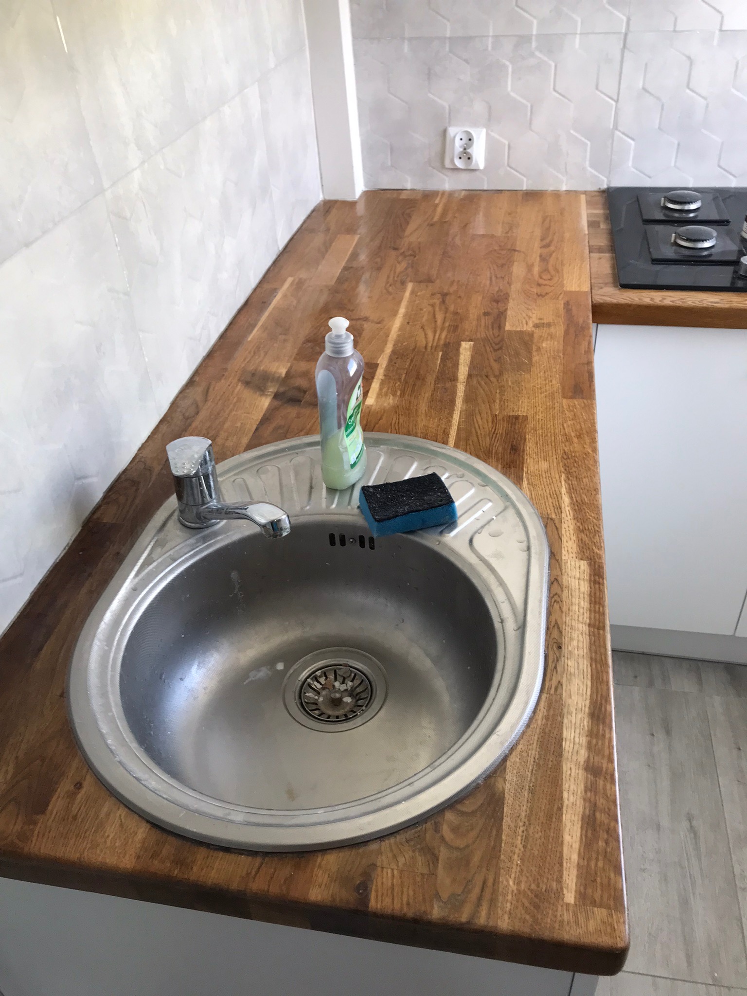 Kuchenny zlew ze stali nierdzewnej wbudowany w drewniany blat, obok płyn do naczyń i gąbka, ściana wyłożona jasnymi płytkami o geometrycznym wzorze.
