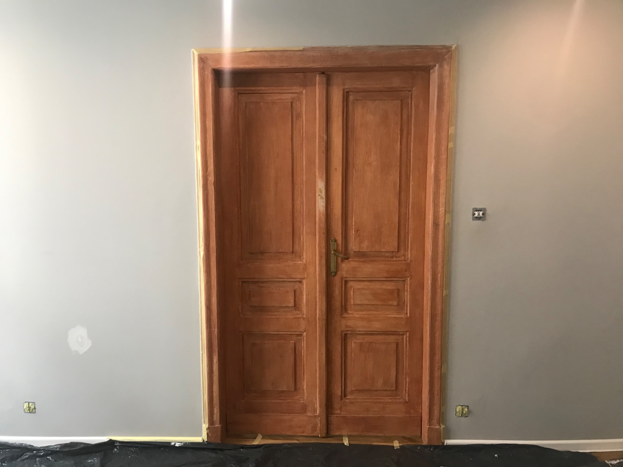 Renowacja dwuskrzydłowych drzwi drewnianych z mosiężną klamką, zabezpieczonych taśmą malarską i folią ochronną na tle szarej ściany z widocznym gniazdkiem elektrycznym i śladami po naprawach.