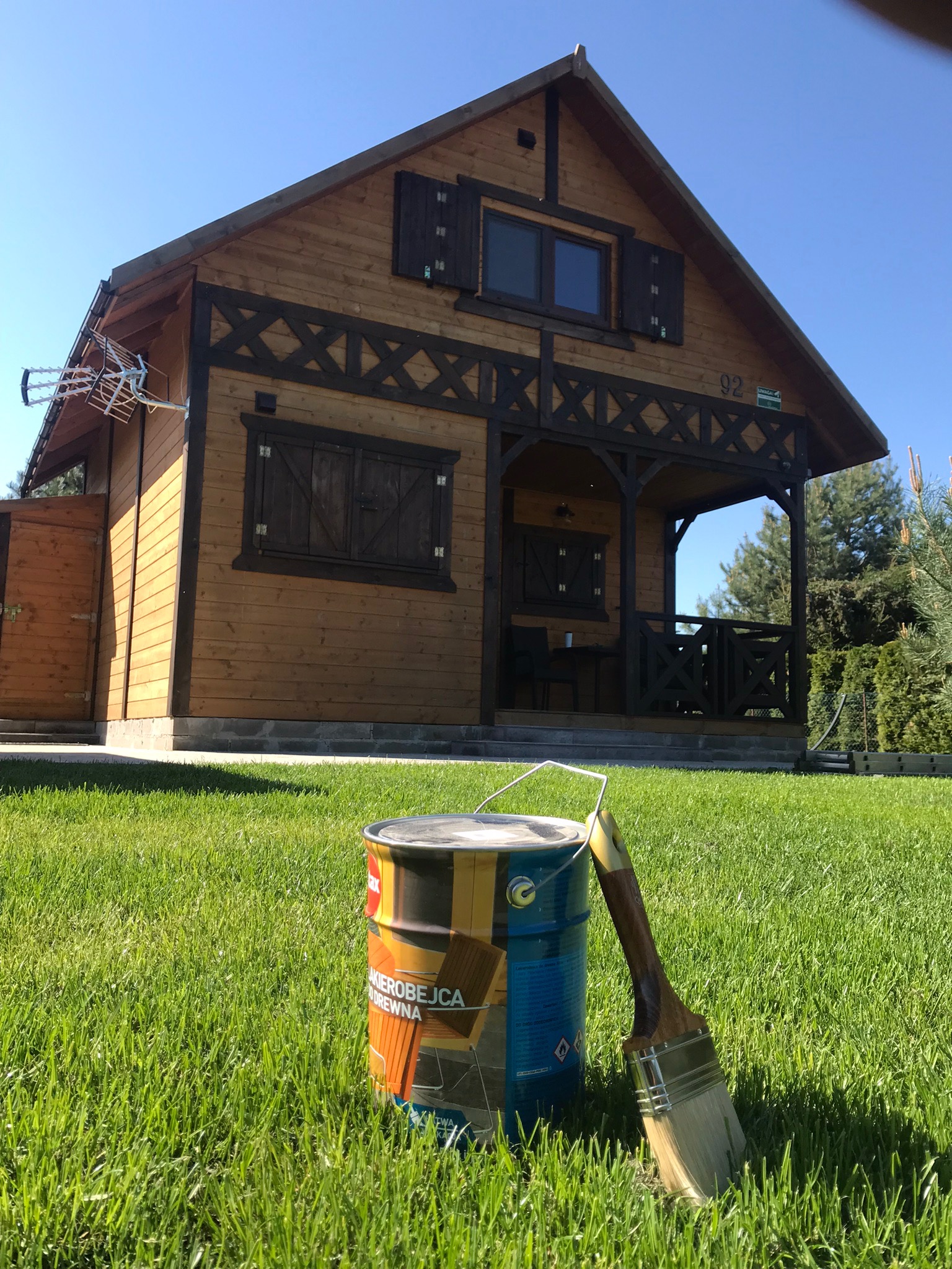 Drewniany domek z ciemnymi okiennicami i balustradą, na pierwszym planie puszka z bejcą do drewna i pędzel na trawie.