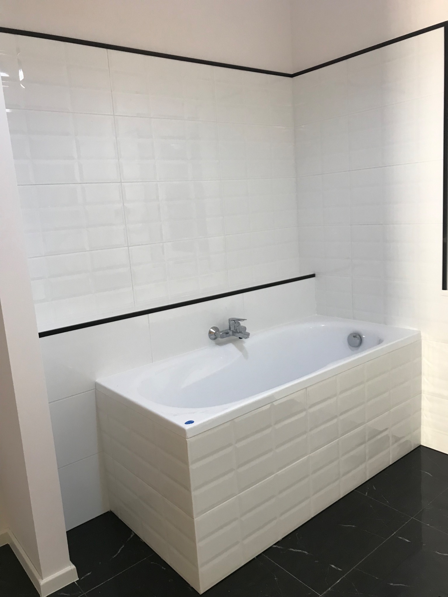 Biała wanna obudowana płytkami imitującymi cegłę, z chromowaną baterią, na tle białych płytek ściennych z czarnym akcentem, oraz czarnej podłogi z marmurkowym wzorem.