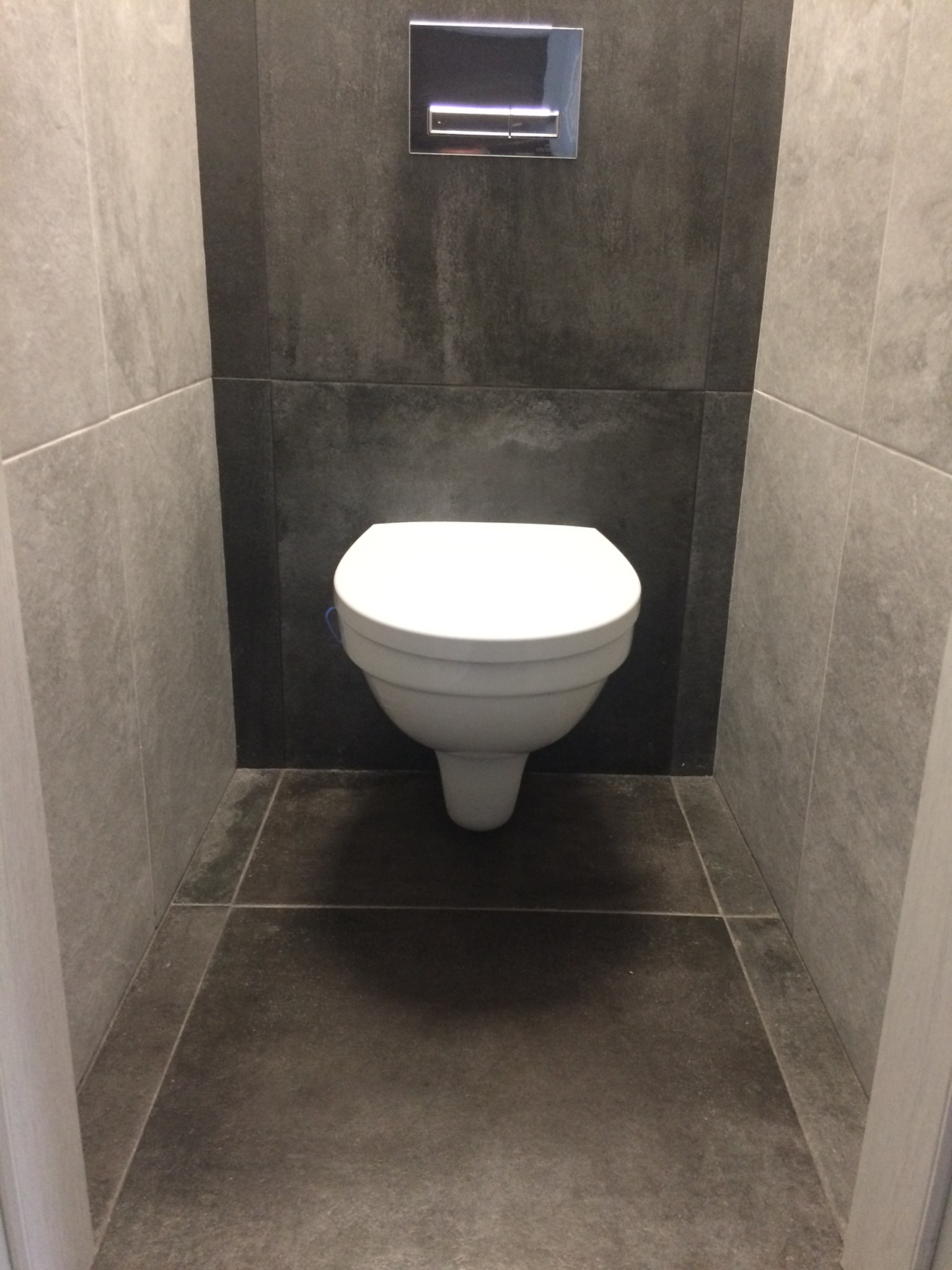 Podwieszana toaleta w nowoczesnej łazience z szarymi płytkami na ścianach i podłodze, widoczny chromowany przycisk spłukiwania.