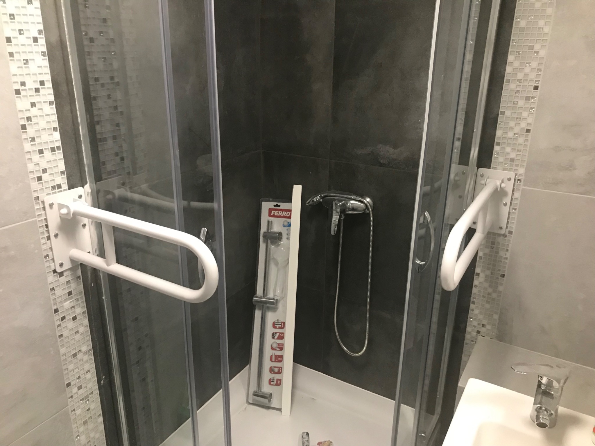 Kabina prysznicowa z zamontowanymi uchwytami dla osób niepełnosprawnych, widoczny również nowy drążek prysznicowy w opakowaniu marki Ferro.