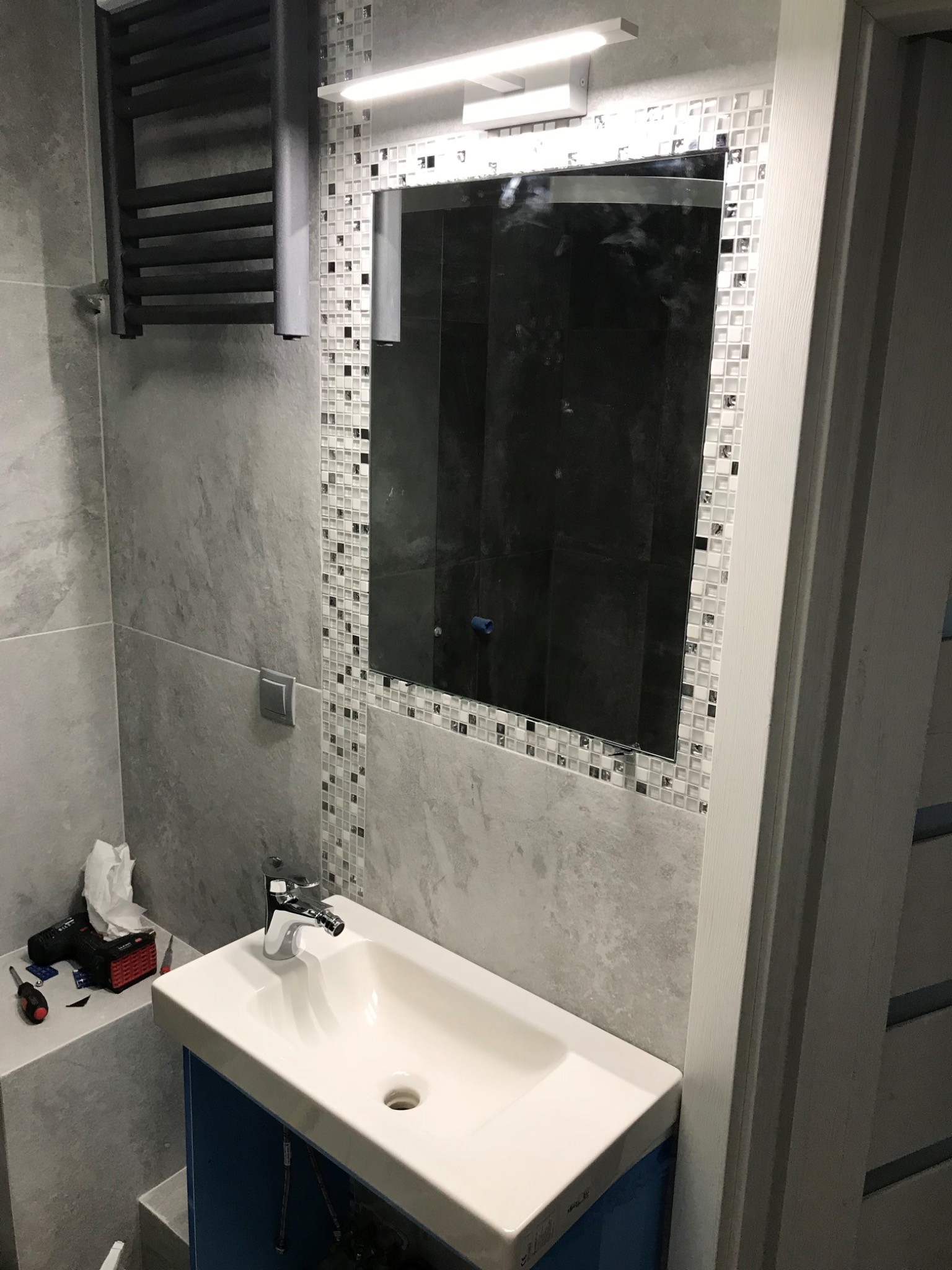 Szara łazienka z umywalką na szafce, lustrem z mozaikową ramą i grzejnikiem drabinkowym; w tle widoczne narzędzia.