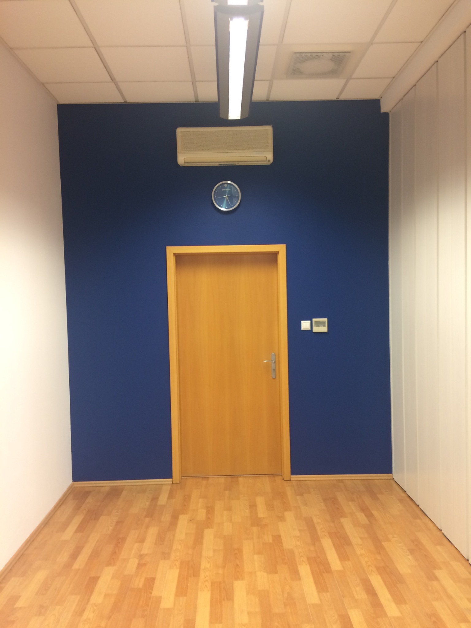Wnętrze biura: drewniane drzwi w ościeżnicy, zegar i klimatyzator na granatowej ścianie, panele podłogowe, oświetlenie sufitowe.