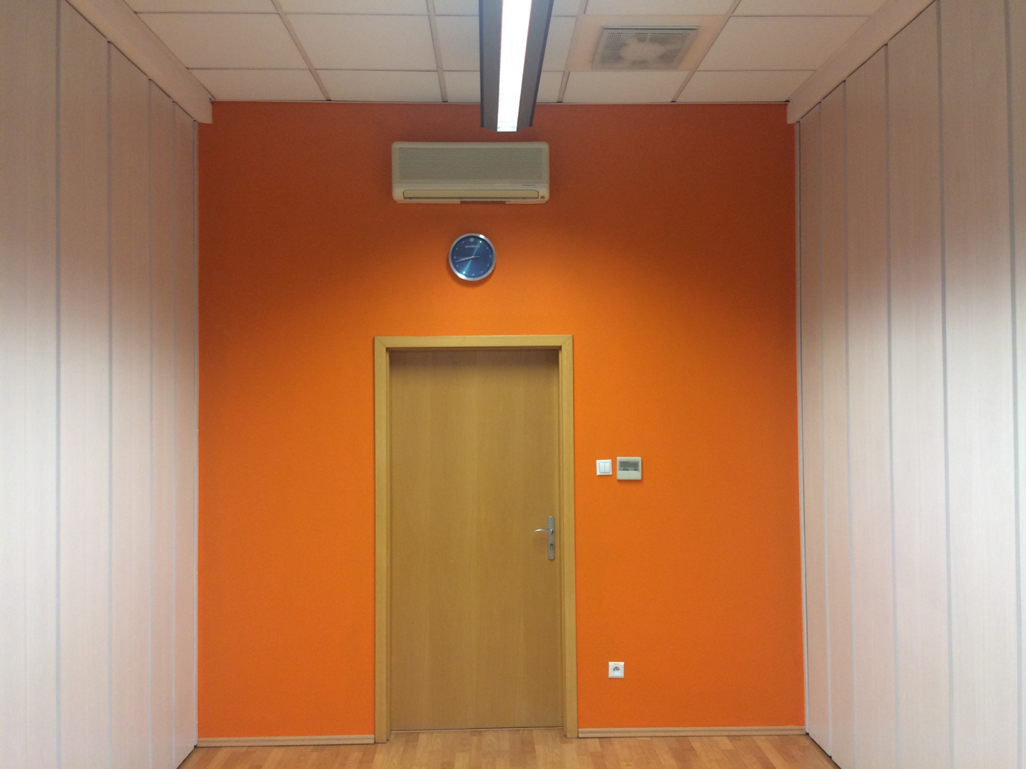 Biuro po remoncie: pomarańczowa ściana z drewnianą ościeżnicą i drzwiami, klimatyzacja, zegar ścienny, białe panele na ścianach bocznych, parkiet.