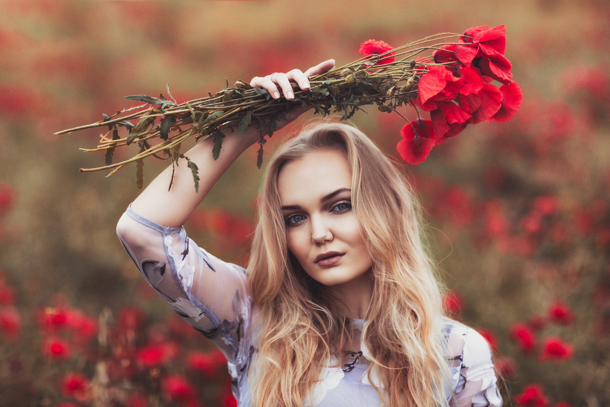 Portret młodej kobiety z blond włosami trzymającej bukiet czerwonych maków na tle pola makowego, ubrana w bluzkę w kwiaty.