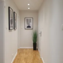 Jasny korytarz z drewnianą podłogą, ozdobiony minimalistycznymi obrazami w czarnych ramkach i wysoką rośliną w czarnej donicy.