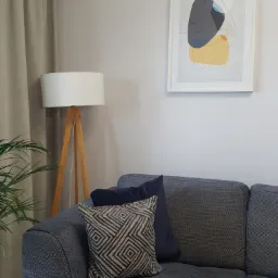 Fragment pokoju z szarą sofą, poduszkami w geometryczne wzory, lampą stojącą z drewnianym trójnogiem i abstrakcyjnym obrazem w białej ramie na ścianie.