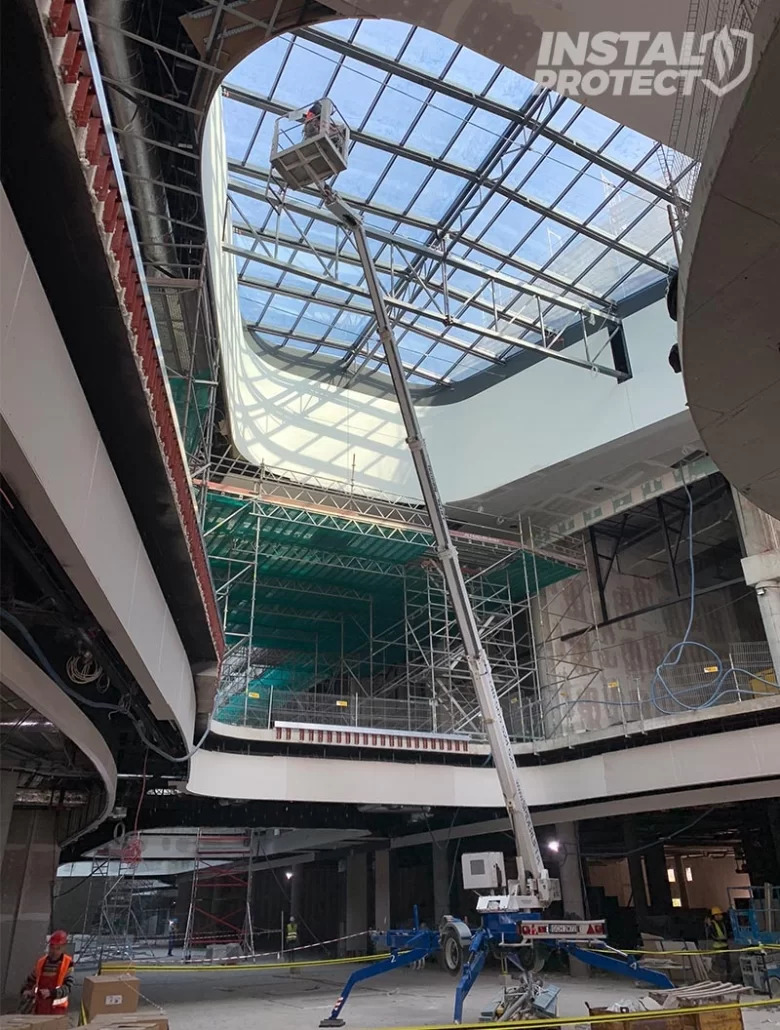 Wnętrze budynku w trakcie budowy z widoczną platformą podnośnikową i pracownikiem przy szklanym dachu, zabezpieczonym siatkami ochronnymi.