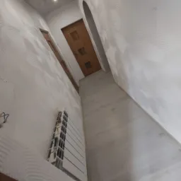 Korytarz z dekoracyjną białą ścianą z wzorem koncentrycznych okręgów, drewniane drzwi w tle, grzejnik w trakcie montażu z wystającymi kablami.