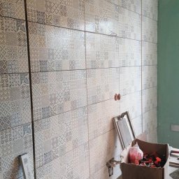 TOM-BUD Tomasz Mielczarek - Ściana łazienki wyłożona patchworkowymi płytkami ceramicznymi w odcieniach szarości i beżu, widoczne narzędzia glazurnicze i karton z klinami do płytek na drewnianym stole.