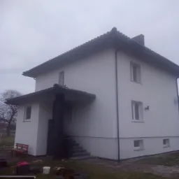 Dwukondygnacyjny dom z jasną elewacją po renowacji, widoczne pozostałości materiałów budowlanych i drewniana ławka przed wejściem.