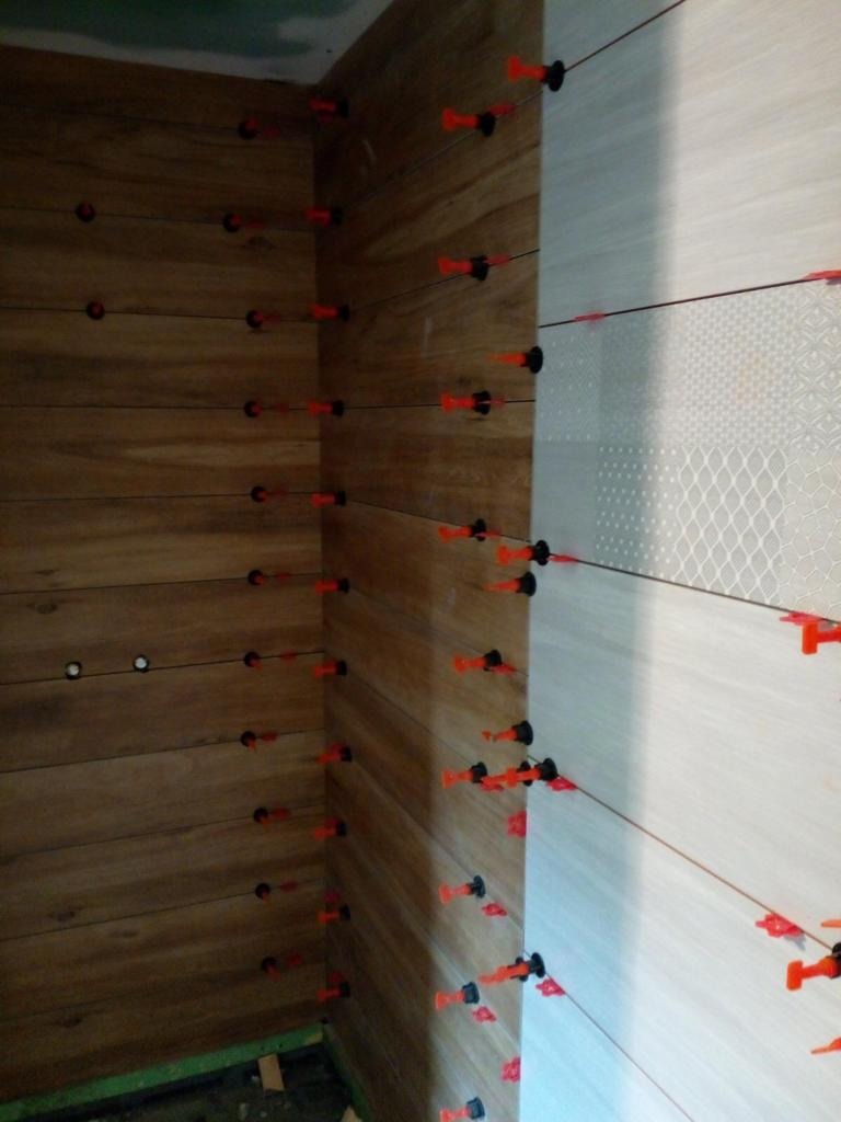 Układanie płytek imitujących drewno na ścianie z użyciem systemu poziomowania z pomarańczowymi klipsami i czarnymi klinami, widoczny narożnik i fragment ściany z jasnymi płytkami z delikatnym wzorem.